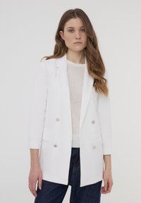 Biała dwurzędowa marynarka z dwoma kieszeniami z przodu. Wykonana z gładkiego materiału, z metalowymi guzikami i nowoczesnym, dopasowanym krojem.