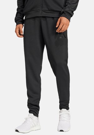 Homme portant un pantalon de jogging Adidas noir et des chaussures de sport blanches, debout, mains visibles et bas du torse sur fond blanc.