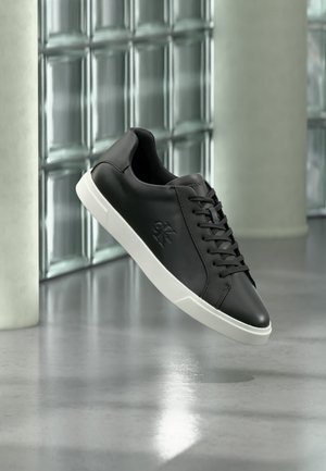 Μαύρο sneaker χαμηλής κοπής με λευκή σόλα, κορδόνια και ανάγλυφο λογότυπο, εμφανίζεται με φόντο από γυάλινες πλάκες και κολώνες από σκυρόδεμα.