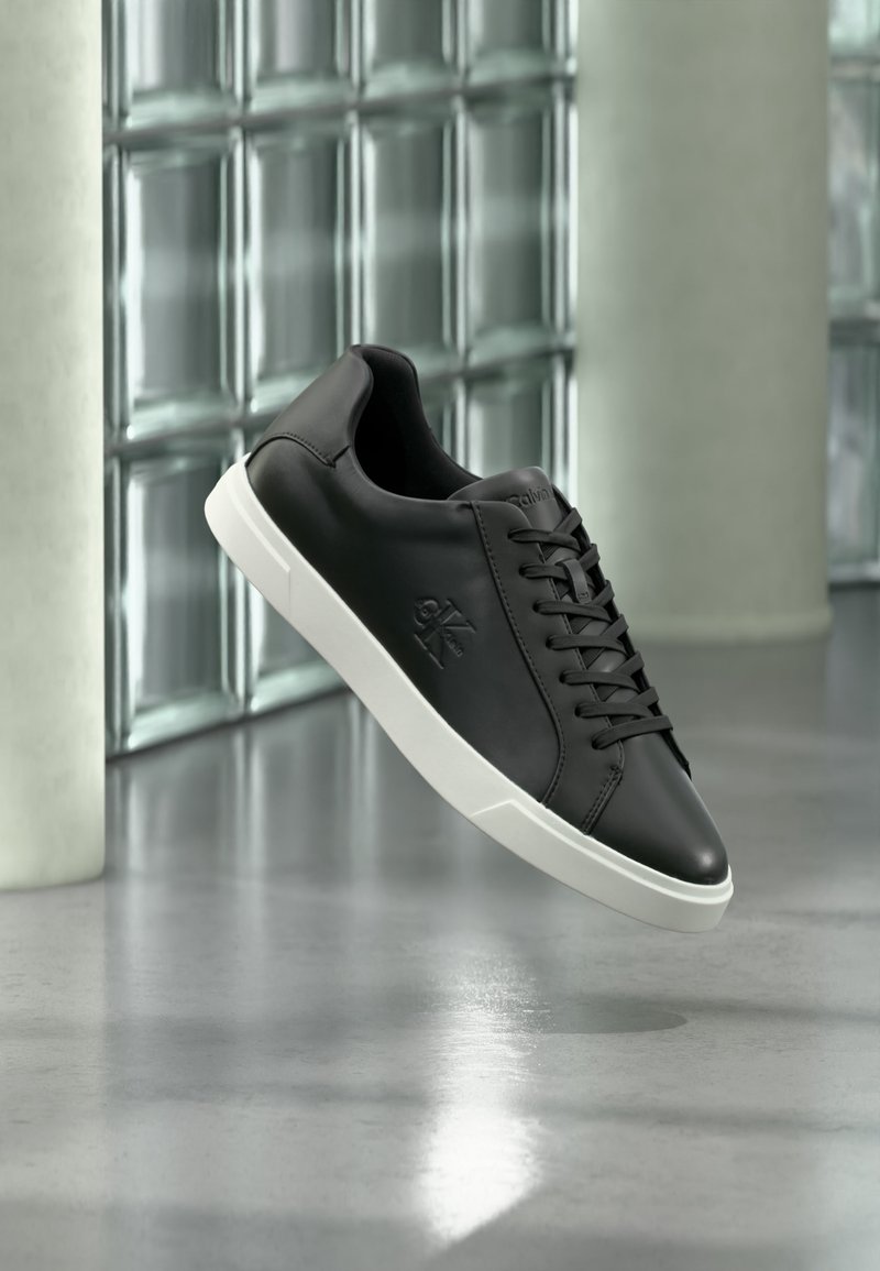 Μαύρο sneaker χαμηλής κοπής με λευκή σόλα, κορδόνια και ανάγλυφο λογότυπο, εμφανίζεται με φόντο από γυάλινες πλάκες και κολώνες από σκυρόδεμα.