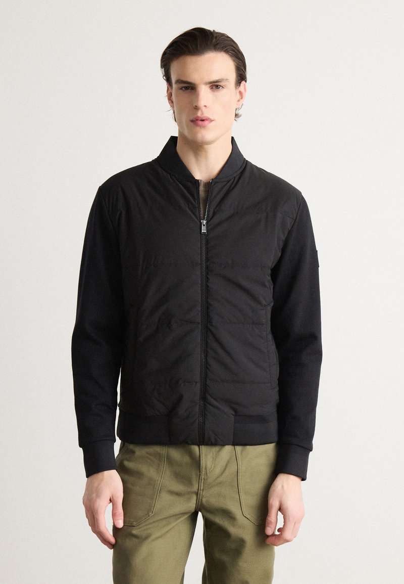 Veste bomber noire avec un devant en nylon matelassé, des poignets côtelés et une fermeture éclair. Le matériau est lisse avec une finition mate.