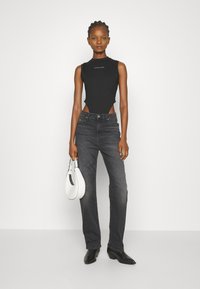 Monos de tirantes negro sin mangas con detalle de corte, combinados con jeans de mezclilla oscuros de tiro alto y botas negras hasta el tobillo, sosteniendo un bolso blanco.