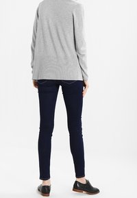 Šedý svetr s dlouhým rukávem a žebrovaným lemem, v kombinaci s tmavě modrými úzkými džíny a černými šněrovacími botami. Pohled zezadu zdůrazňuje střih outfitu.