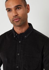 Sort kordfløjs skjorte med knapper og krave, forsynet med en brystlomme og tekstureret stof. Synlige knapper i kontrastfarve.