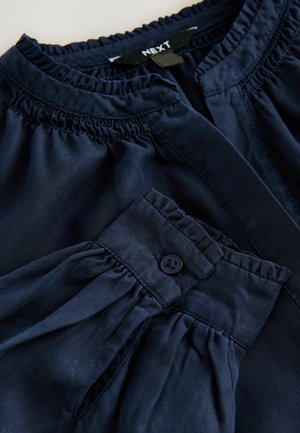 Navyblauwe blouse met geplooide halslijn en knoopsluiting bij de manchetten, gemaakt van zachte stof met een gladde textuur en een taps toelopende vorm.