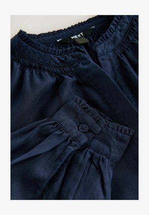 Blouse bleu marine avec encolure froncée et poignet à boutons, fabriquée en tissu doux avec une texture lisse et une forme cintrée.