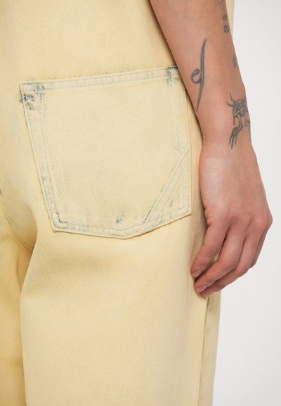 Κοντινό πλάνο ενός χεριού με τατουάζ, που ακουμπά δίπλα στην πίσω τσέπη ενός κίτρινου τζιν παντελονιού με διακριτικά σημάδια φθοράς.