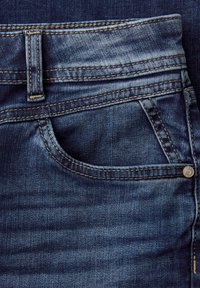 Mørkeblå denimjeanslomme med synlige gule sømme, bæltestrop og sølvnitteknap på tekstureret stof.