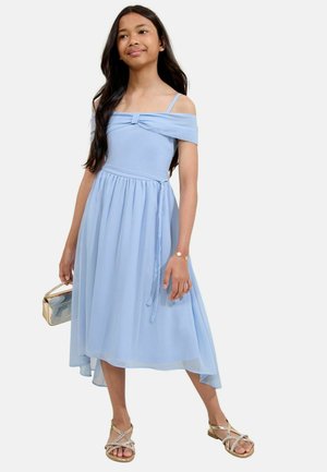 Mädchen in einem hellblauen schulterfreien Kleid, das eine goldene Handtasche hält, goldene Sandalen trägt und ein sternförmiges Armband trägt, steht vor einem schlichten Hintergrund.