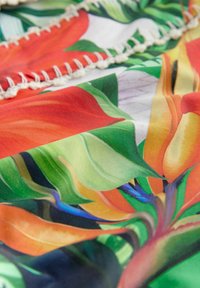 Kleurrijke stof met grote bloemenpatronen in rood, oranje, groen en blauw. Zachte textuur met een subtiele glans en decoratieve gestikte randen.