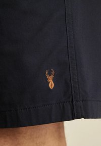 Pantalones cortos azul marino hechos de una tela suave, con un pequeño logotipo de ciervo bordado en marrón claro cerca del dobladillo, y detalles de costura bien hechos.