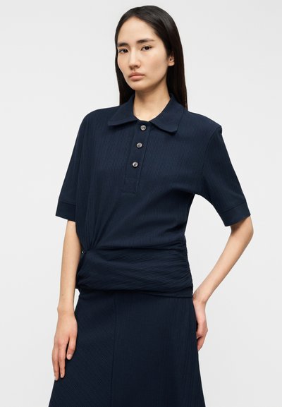 Πολύ μπλε πόλο μπλουζάκι με ριμπ ύφασμα και κοντά μανίκια, με γιακά και τρεις κουμπότρυπες, σχεδιασμένο με στριφτή λεπτομέρεια στη μέση.