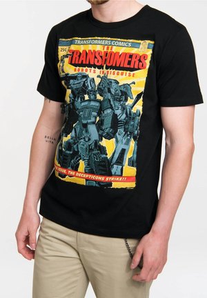 Mann trägt schwarzes T-Shirt mit Vintage-Style "Transformers" Comic-Cover, das Roboter und fetten roten Text auf gelbem Hintergrund zeigt.