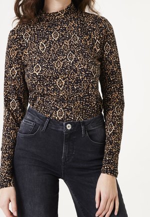 Nainen, jolla on päällään pitkähihainen, musta ja ruskeakuvioinen poolopaita, joka on sujautettu korkeavyötäröisten mustien farkkujen sisään valkoista taustaa vasten.