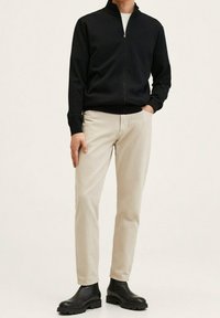 Chaqueta negra con cremallera, pantalones beige claros y botas negras. La chaqueta tiene un cuello alzado y puños acanalados. Los pantalones son de algodón ajustados.