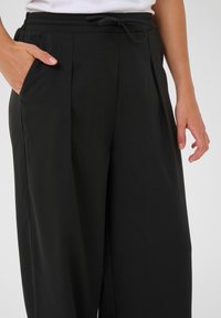 Pantalons larges noirs en matériau léger. Comprend une taille à cordon de serrage et des poches latérales. Texture lisse avec des plis.