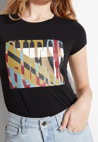 Mustat t-paita, jossa on paljeteilla koristeltu "GUESS"-logo, joka esittelee monivärisiä kultaisia, punaisia, sinisiä ja valkoisia yksityiskohtia kuviollisella taustalla.
