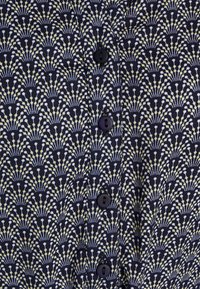 Chemise à boutons avec un fond bleu marine et un motif floral en blanc et jaune. Elle a des boutons noirs et une texture de tissu lisse.