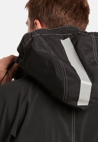 Chaqueta negra con capucha, costuras blancas y una franja reflectante en la parte de atrás. El material parece ligero y resistente al agua.