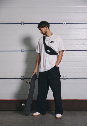 Młody mężczyzna noszący koszulkę Nike SB i torbę przez ramię, trzymający deskorolkę, stoi na tle białych drzwi garażowych.