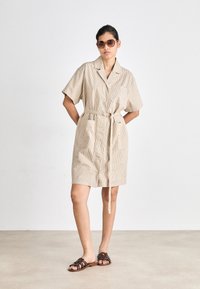 Strikket beige skjortekjole med korte ermer, knappestolpe foran og stoffbelte. Har to frontlommer og en avslappet passform. Brune sandaler.