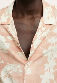 Personne à la peau foncée portant une chemise pêche à boutons avec de grands motifs floraux blancs et un col ouvert.