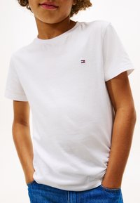 Kind trägt weißes T-Shirt mit kurzen Ärmeln und kleinem Flaggenlogo auf der Brust sowie blaue Jeans, Hände in den Taschen, vor einfarbigem Hintergrund.