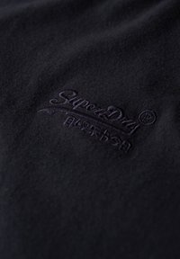 Tela negra, primer plano del logotipo bordado "SuperDry" en púrpura oscuro. Textura suave, patrón minimalista con detalle del logotipo.