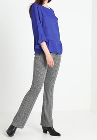 Blusa blu royal con maniche a volant e vestibilità ampia abbinata a pantaloni grigi a fantasia e stivali neri alla caviglia, in piedi su uno sfondo bianco.