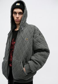 Sudadera acolchada gris con capucha, bolsillos laterales, puños acanalados y un pequeño parche con el logo en el pecho. Textura suave y ajuste relajado.
