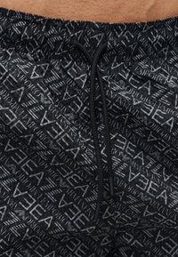 Zwarte shorts met trekkoord en herhalende "Emporio Armani" en "EA7" logo's in grijs, gedragen op de taille met zichtbare huid erboven.