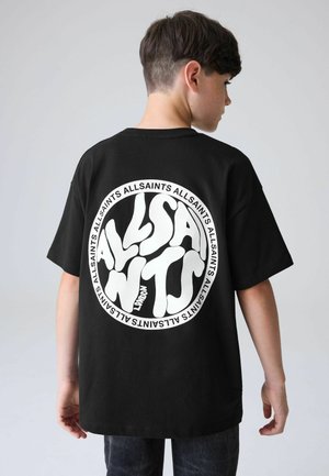Poika, joka seisoo sivuttain, päällään musta t-paita, jossa on suuri valkoinen pyöreä "ALLSAINTS LONDON" -logo selässä, tasaisella taustalla.