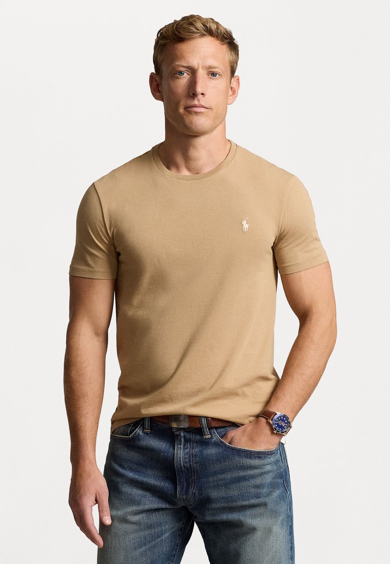 Beige īsām piedurknēm T-krekls ar apaļu kaklu un mazu logotipu uz krūts, apvienots ar ziliem džinsiem un brūnu pulksteni uz rokas.
