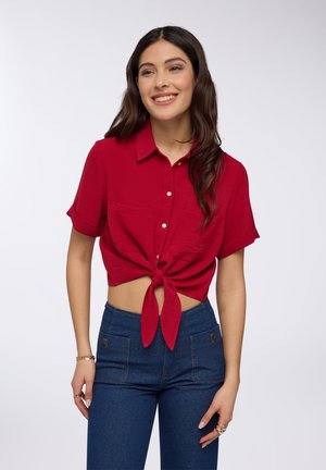 Camicia rossa a maniche corte con bottoni e nodo sul davanti, in tessuto testurizzato, con due tasche sul petto, abbinata a pantaloni in denim blu a vita alta.