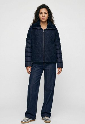Frau steht frontal, trägt eine dunkelblaue strukturierte Jacke mit gesteppte Ärmeln, Jeans mit weitem Bein und silberne Sneaker vor einfarbigem Hintergrund.