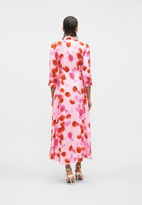 Γυναίκα με πλεξούδες που φοράει ροζ midi φόρεμα με σχέδια από κόκκινα και ροζ πέταλα και λευκά ψηλοτάκουνα σανδάλια, στέκεται γυρισμένη προς τα πίσω.