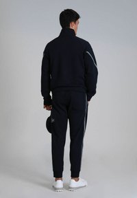 Navy Trainingsanzug mit hohem Kragen und weißen Streifen entlang der Ärmel und Seiten, kombiniert mit weißen Turnschuhen. Das Modell hält einen schwarzen Hut.