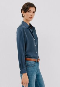 Blusa di seta blu navy con colletto, chiusura frontale con bottoni e maniche lunghe. Abbinata a jeans blu chiaro e una cintura in pelle marrone.