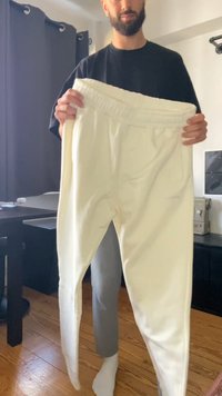 Bele hlače za telovadbo z elastičnim pasom, zoženimi nogavicami, mehko teksturo in minimalnimi podrobnostmi, ki jih drži oseba v črnem majici.