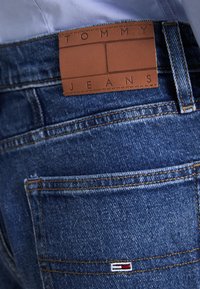 Modré džíny z denimové látky mají hnědou koženou nášivku se nápisem "TOMMY JEANS." Materiál má typickou denimovou strukturu s kontrastním prošíváním.