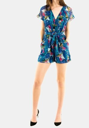 Femme portant une combinaison courte bleue à manches courtes et col en V avec un motif de feuilles tropicales et une ceinture à la taille, associée à des chaussures à talons noirs.