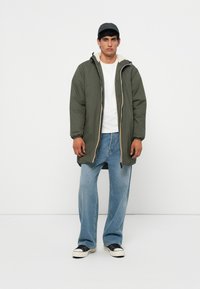 Veste matelassée verte avec un col beige, associée à un t-shirt blanc et un jean large bleu clair. Des baskets noires complètent la tenue.