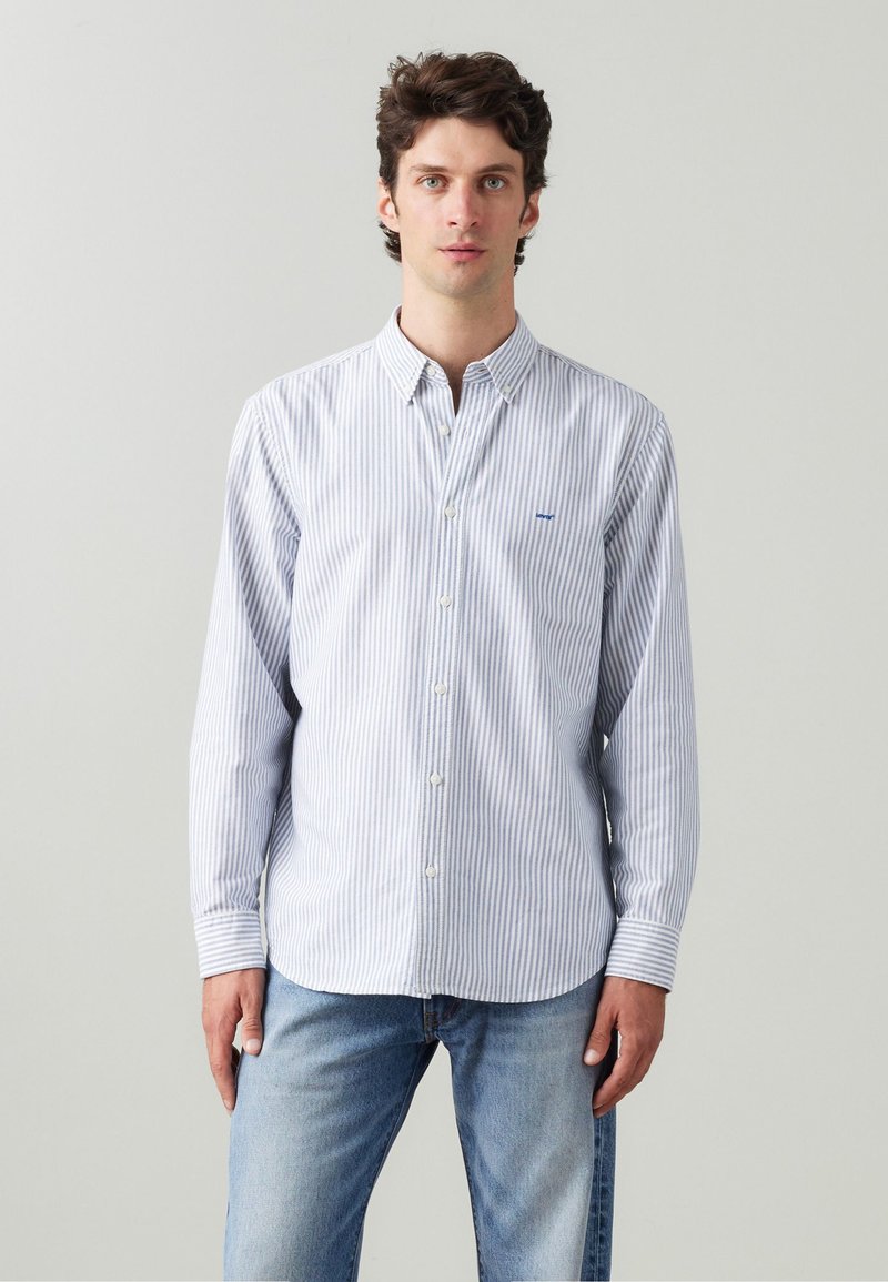 Chemise boutonnée rayée bleu clair et blanche avec col et manches longues. Dotée d'un petit logo bleu sur la poitrine. Tissu en coton.