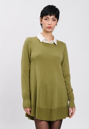 Femme aux cheveux courts noirs portant une robe tunique manches longues vert olive sur une chemise blanche à col, debout devant un fond uni.