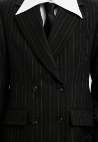 Črn pinstripes dvoredni suknjič z kontrastnim belim ovratnikom, črnimi gumbi in žepom na prsih; tkanina se zdi teksturirana.