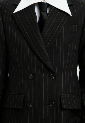 Chaqueta de traje de dos botones a rayas negras con un cuello blanco en contraste, botones negros y un bolsillo en el pecho; la tela parece texturada.