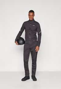 Dunkelgraue Radfahrjacke und -hose aus leichtem Material mit reflektierenden Akzenten. Das Modell hält einen schwarzen Helm.