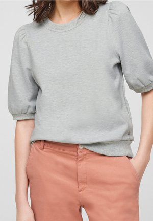 Vrouw draagt een lichtgrijs kort topje met pofmouwen, lichtjes ingestopt in een hoog getailleerde, stoffig roze broek met handen in de zakken.