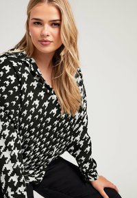 Camisa de patrón houndstooth en blanco y negro con cuello abotonado. Mangas largas y textura de tela suave. Modelo sentado con pantalones negros.