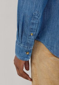 Blauwe denimshirt mouwen met gouden knoopdetails en gestikte manchetten, in combinatie met de textuur van beige corduroy stof die daaronder zichtbaar is.